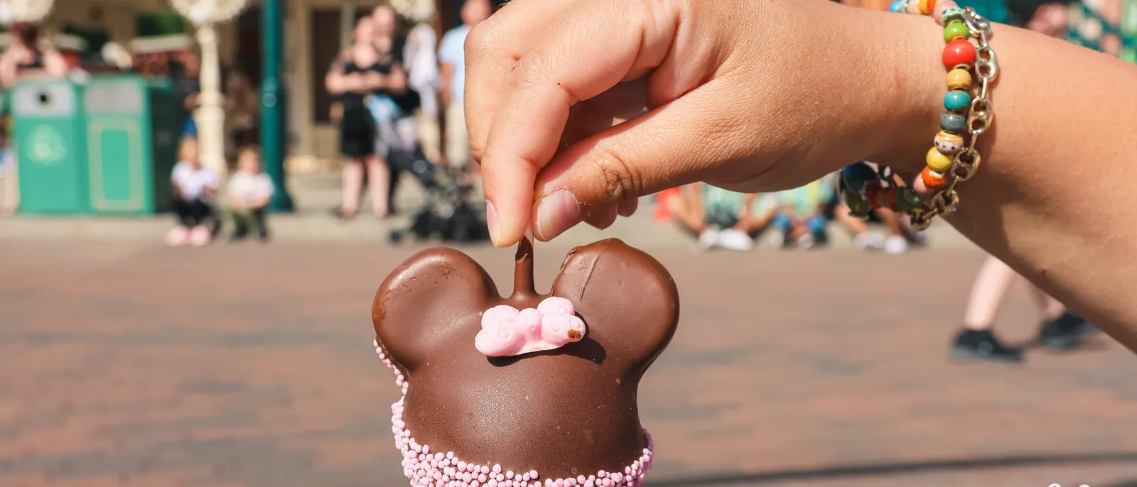 Main tenant un cake pop en forme de Mickey enrobé de chocolat et de billes roses, devant Main Street.