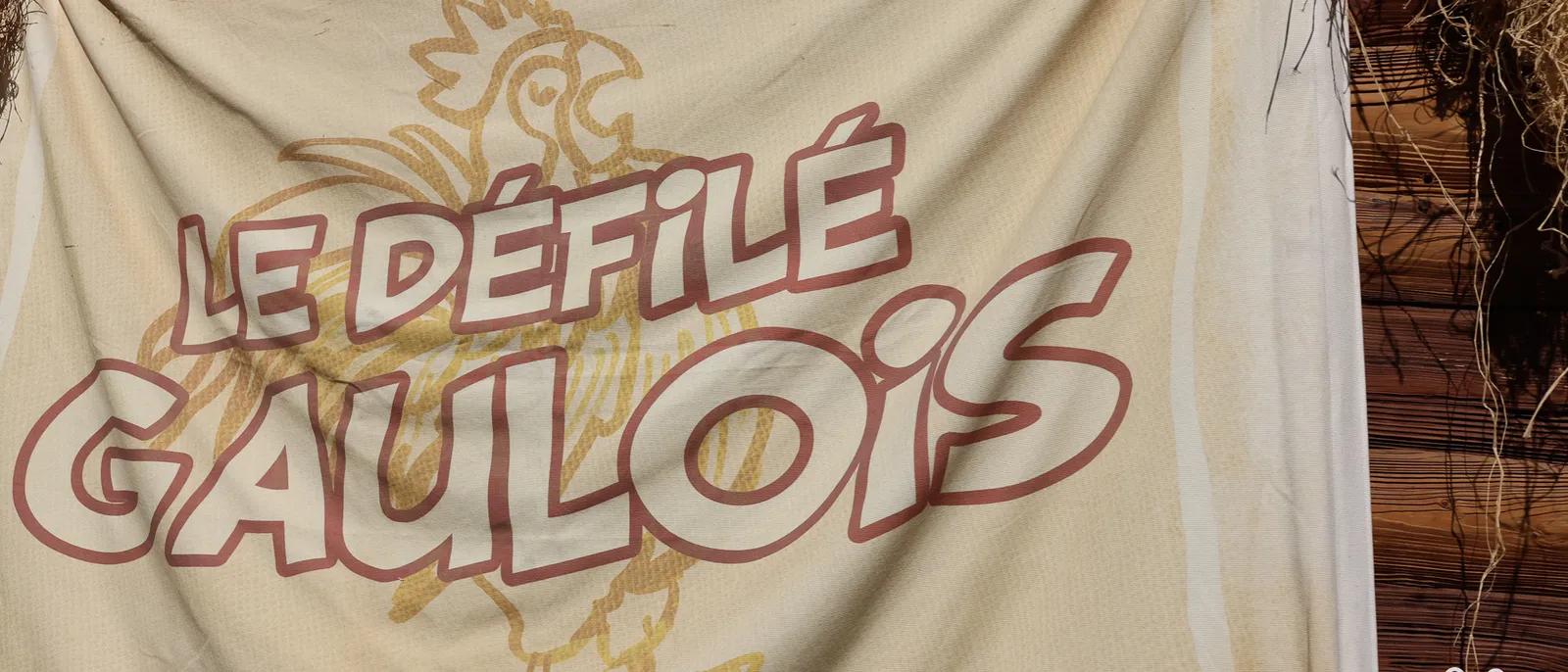 Toile beige tendue peinte du titre Le Défilé Gaulois en lettres blanches cerclées de rouge, coq jaune en fond