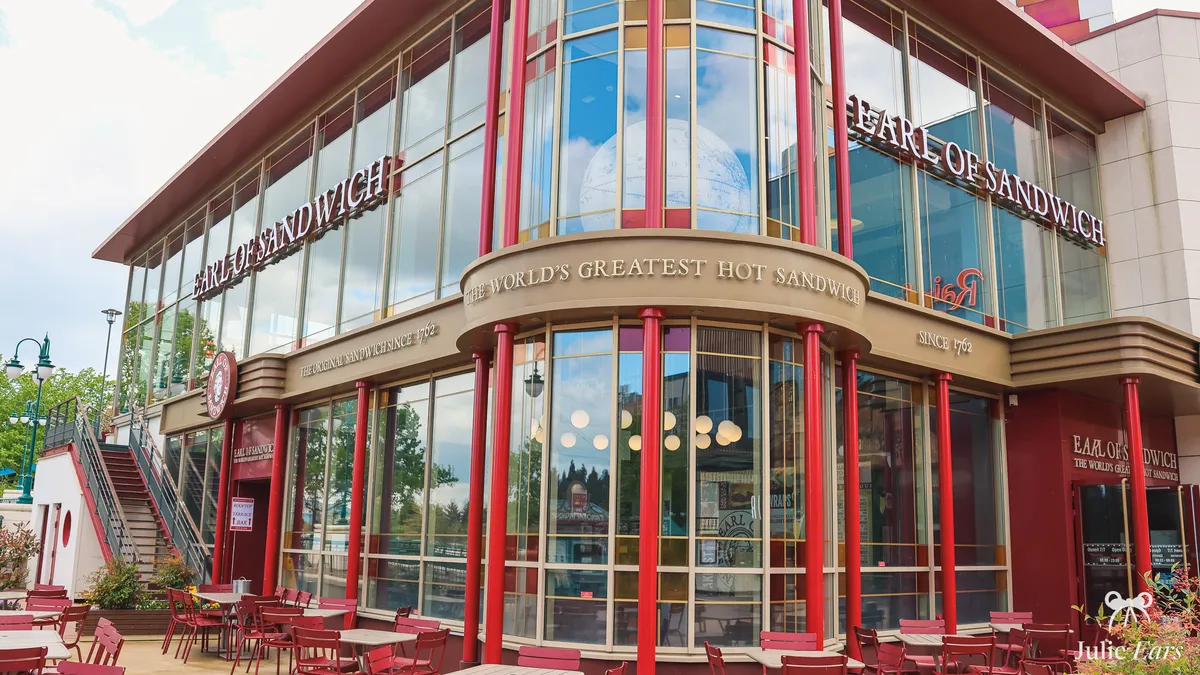 Façade vitrée d’Earl of Sandwich au Disney Village, avec ses colonnes rouges, l’enseigne "The World’s Greatest Hot Sandwich" et sa terrasse aux chaises rouges sous un ciel voilé
