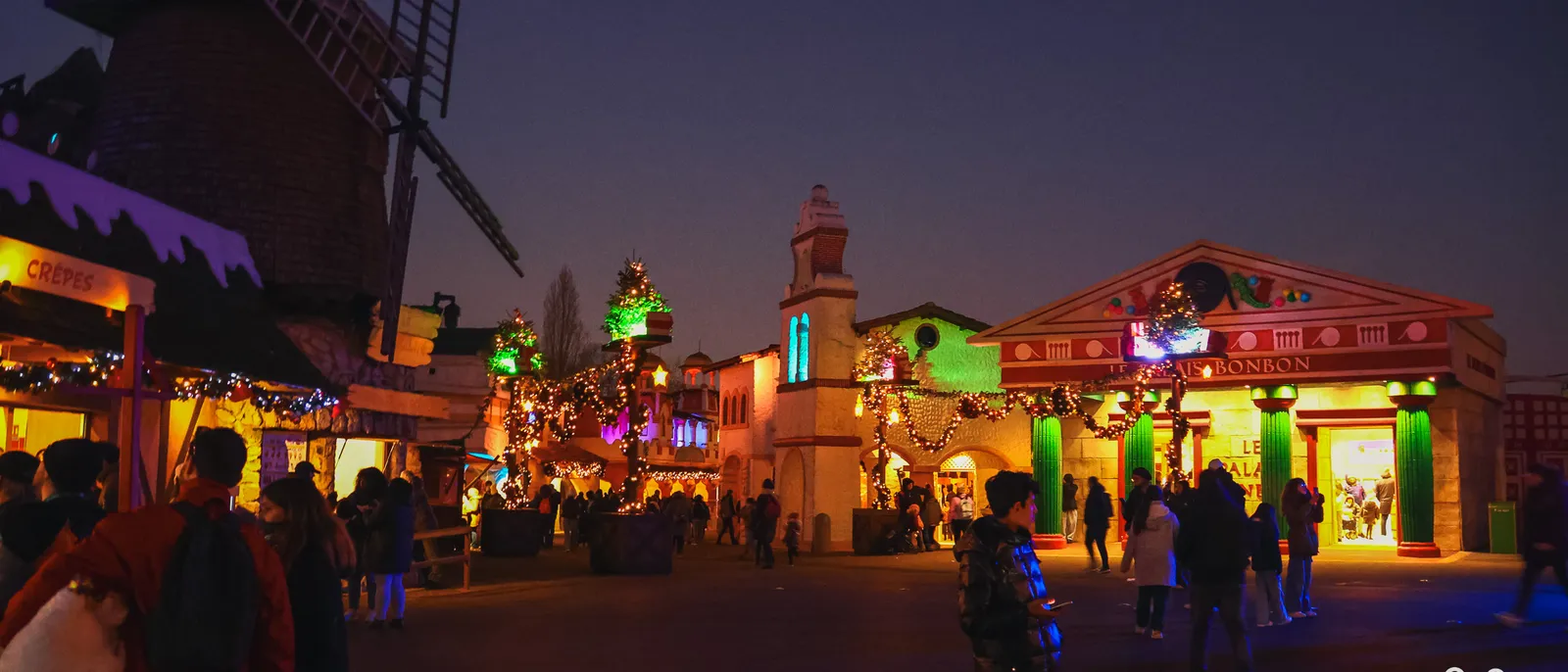 Allée du parc illuminée à la nuit tombée pour Noël, moulin à vent et façades ornées de guirlandes lumineuses, visiteurs de dos