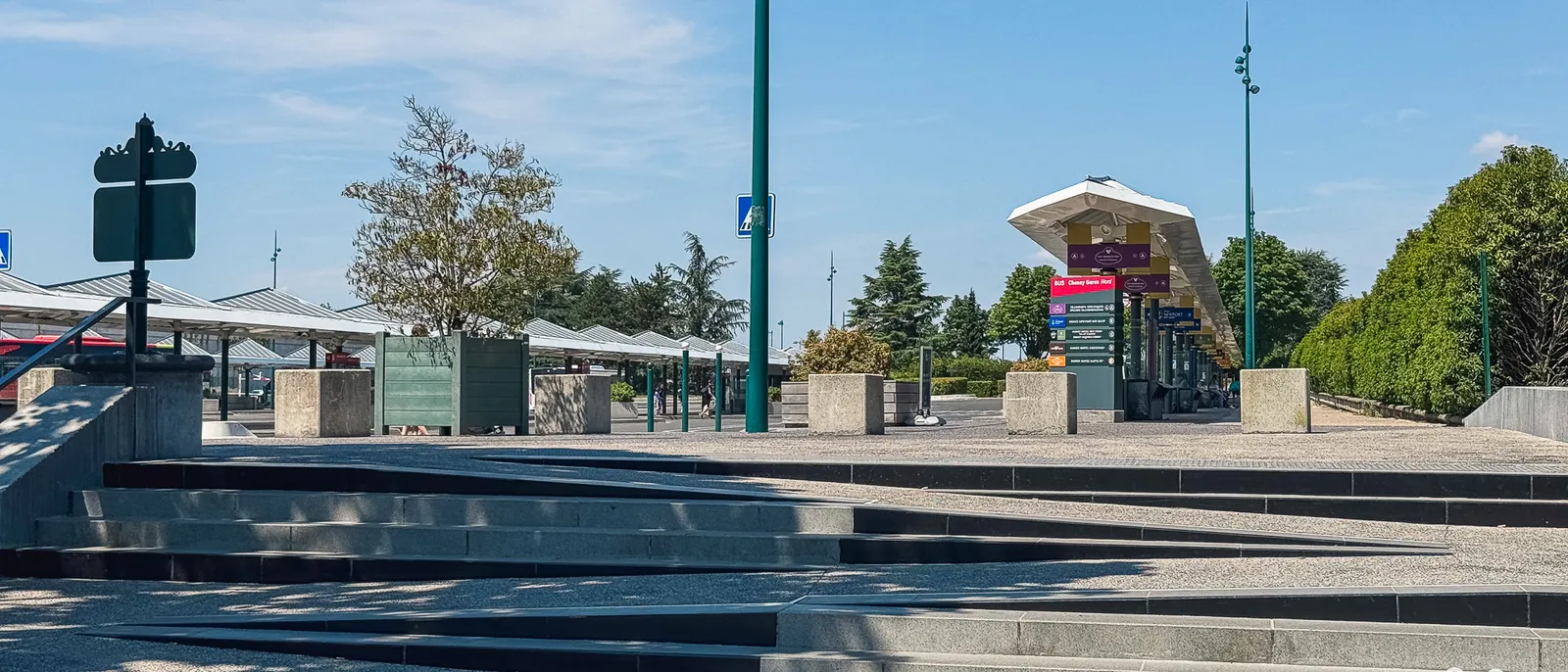 Esplanade de la gare routière de Disneyland Paris avec ses quais couverts et un kiosque, sous un ciel bleu.
