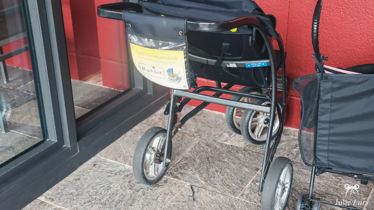 Disneyland Paris hire pushchair parked against a red wall: black metal frame, large grey wheels and a clear pouch on the handlebar for the name