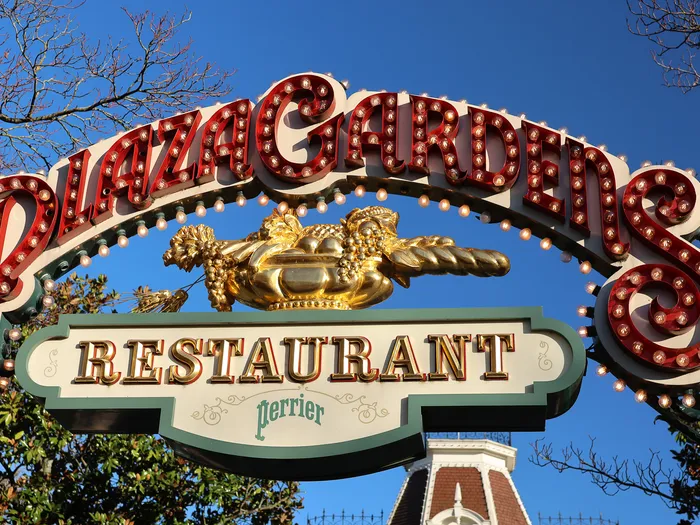 Enseigne lumineuse rouge et or du Plaza Gardens Restaurant sous un ciel bleu, sur Main Street USA.