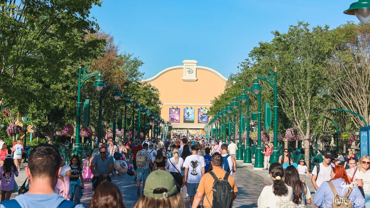 Crowds on Adventure Way at Disney Adventure World, Disneyland Paris, lined with trees and green lampposts, facing a building with the Disney logo