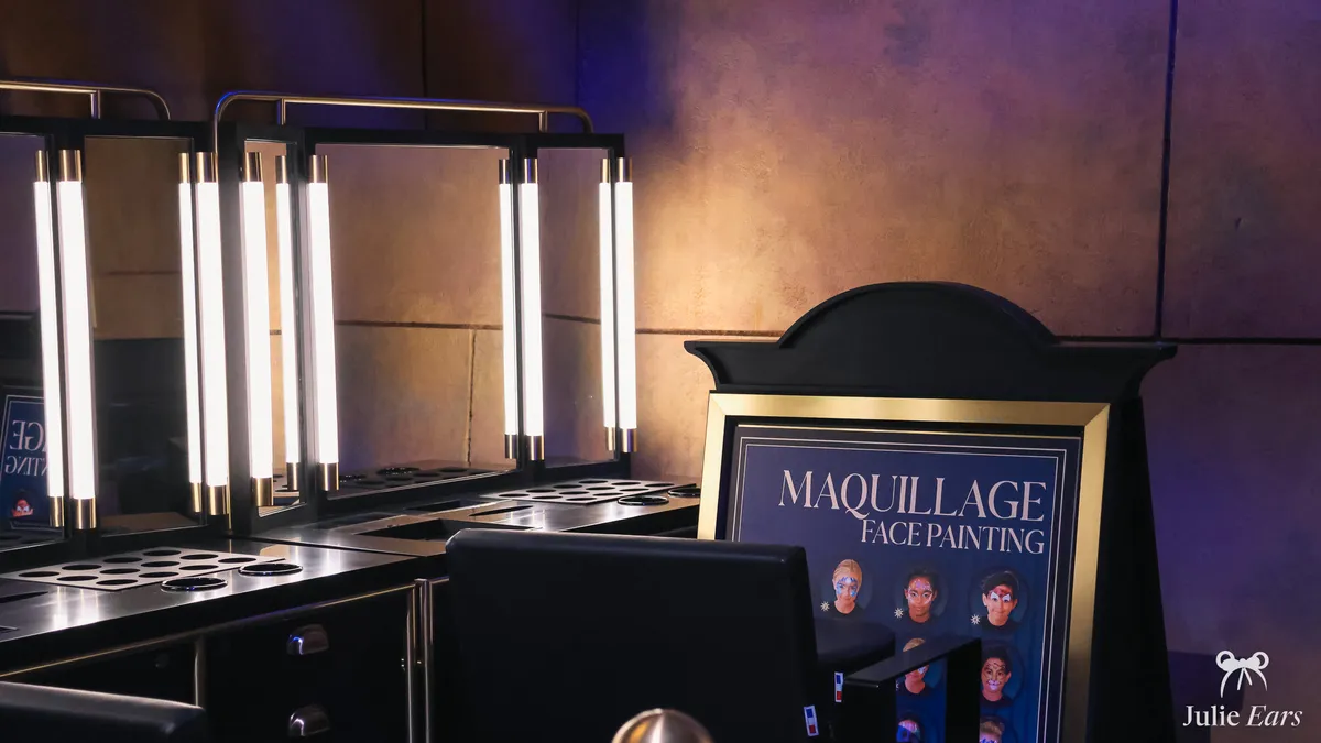 Stand de maquillage (face painting) de Disneyland Paris : miroirs lumineux, palettes et panneau présentant les modèles proposés.
