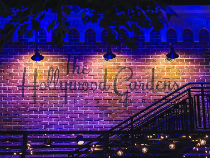 Mur de briques éclairé de violet portant l’enseigne The Hollywood Gardens, avec escalier et guirlandes lumineuses.