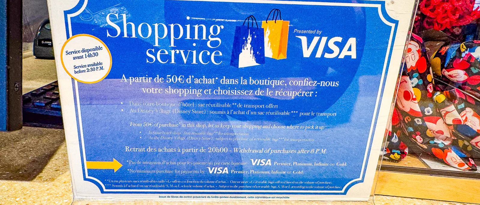 Panneau bleu Shopping service presented by Visa posé sur un comptoir de boutique Disneyland Paris.
