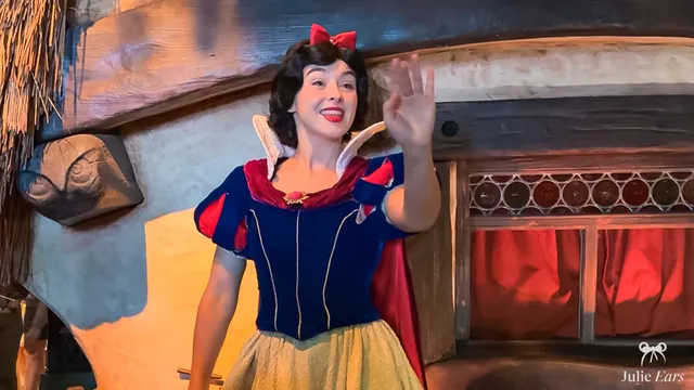 La princesse Blanche-Neige, nœud rouge dans les cheveux et robe bleue et jaune, salue devant la maison des Sept Nains à Disneyland Paris.