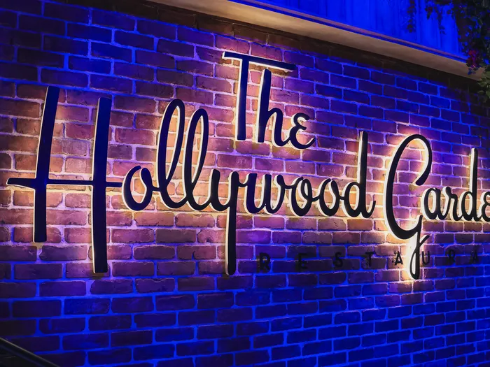 Enseigne cursive lumineuse The Hollywood Gardens sur un mur de briques éclairé de bleu