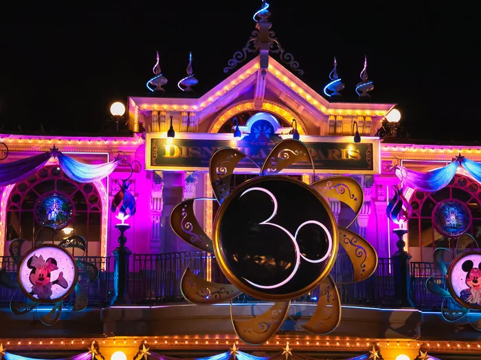 Façade de la gare Disneyland Paris éclairée en rose et bleu la nuit, avec le logo des 30 ans