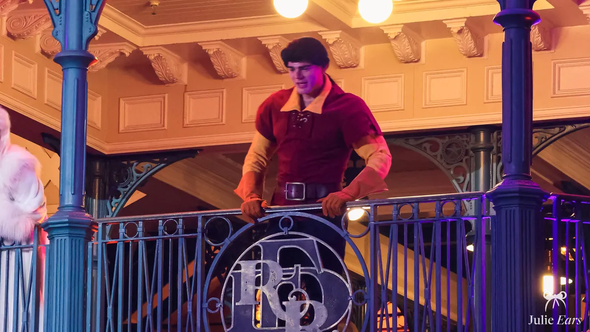 Gaston, in a red shirt and with his imposing build, leaning on a railing during an appearance at Disneyland Paris.