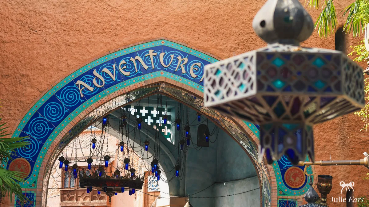 Blue and turquoise mosaic arch bearing the Adventureland name at Disneyland Paris, blue glass chandeliers hanging under the vault and a blurred oriental lantern in the foreground, in the sunshine