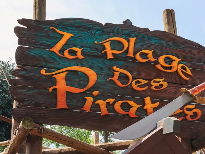 Enseigne de bois gravée « La Plage des Pirates » en lettres orange, ornée d’un sabre, à l’entrée de l’aire de jeux.