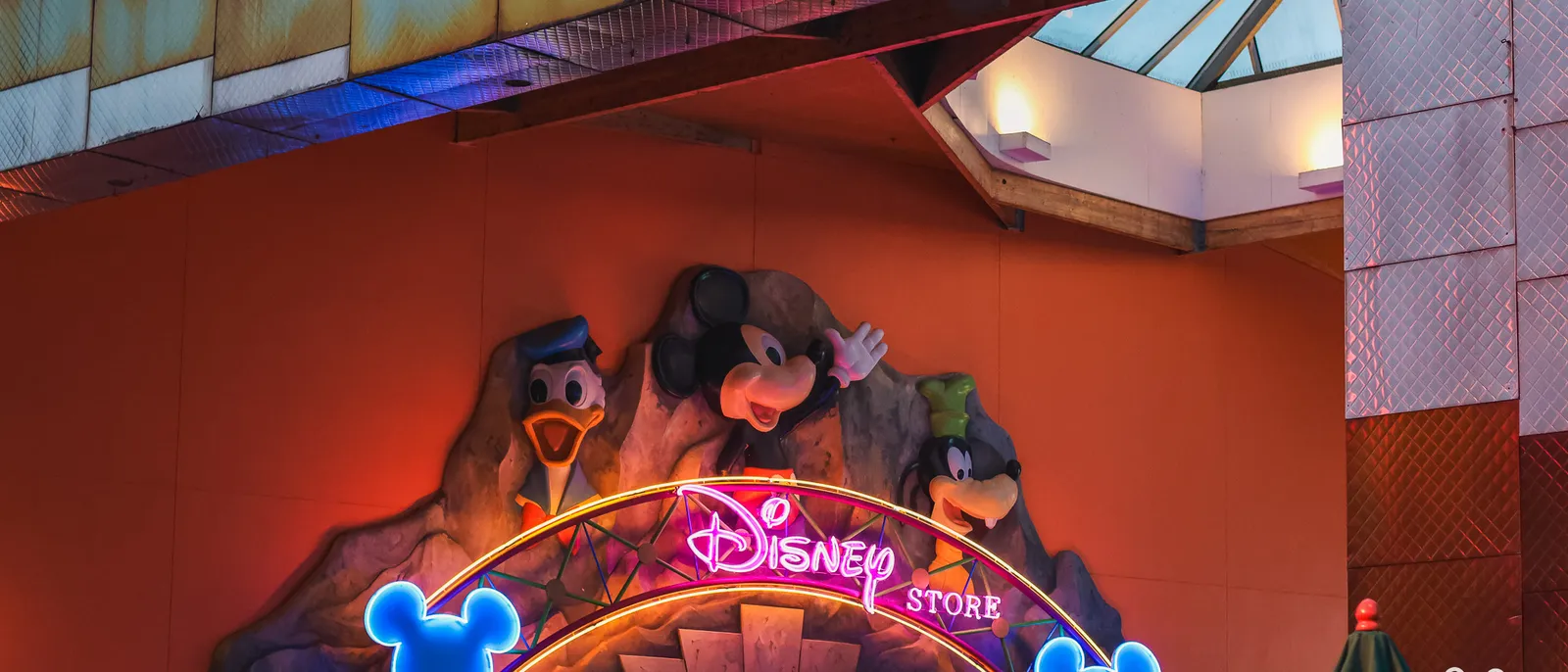 Façade du Disney Store au crépuscule, enseigne néon rose et visages sculptés de Mickey, Donald et Dingo.