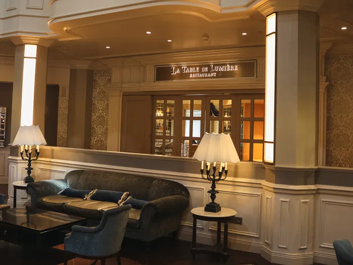 Salon feutré avec canapé et lampes devant l’entrée éclairée du restaurant La Table de Lumière au Disneyland Hotel.