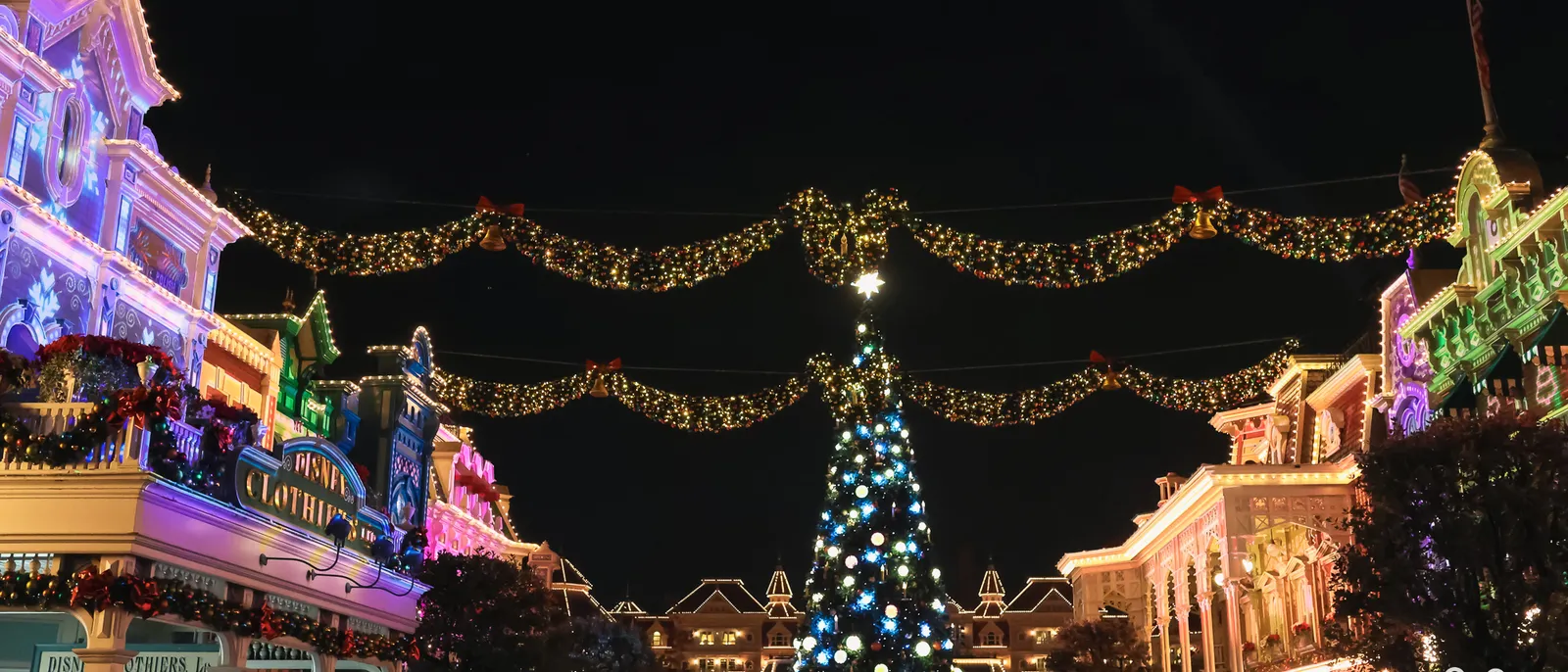 Grand sapin de Noël illuminé au bout de Main Street la nuit, façades et guirlandes lumineuses de part et d’autre.