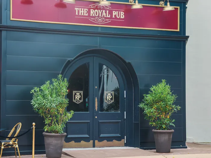 Porte cintrée et enseigne dorée de l’entrée du Royal Pub au Disney Village, avec plantes et chaises bistrot.
