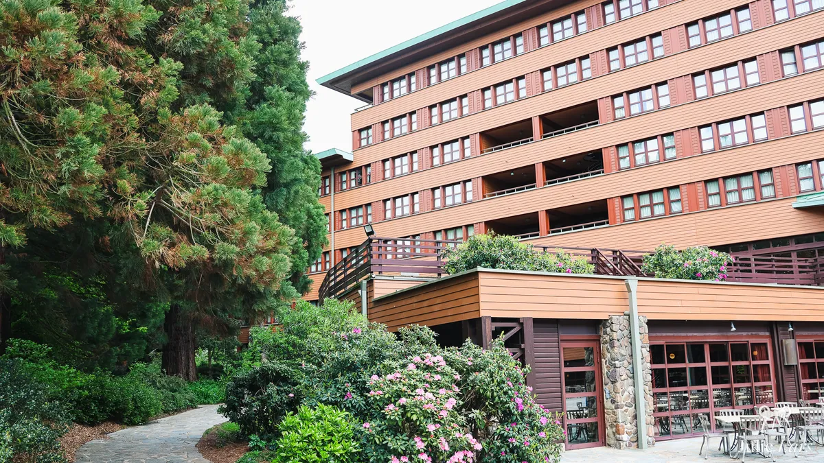 Disney Sequoia Lodge at Disneyland Paris seen from a garden path, with tall conifers, pink rhododendrons in bloom and a terrace with metal tables