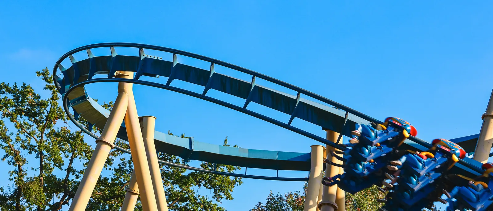 Train du coaster OzIris suspendu sous ses rails bleus, piliers beiges et cimes d’arbres sous un ciel dégagé