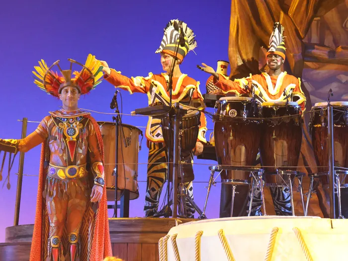 Un chanteur et deux percussionnistes aux congas en costumes colorés sur la scène du spectacle Le Roi Lion