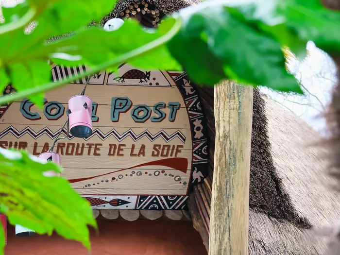 Enseigne peinte du Cool Post « sur la route de la soif », sous un toit de chaume et entourée de feuillages.