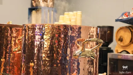Large steaming copper pot of mulled wine, with its tap and the cups ready, at the food festival chalet.