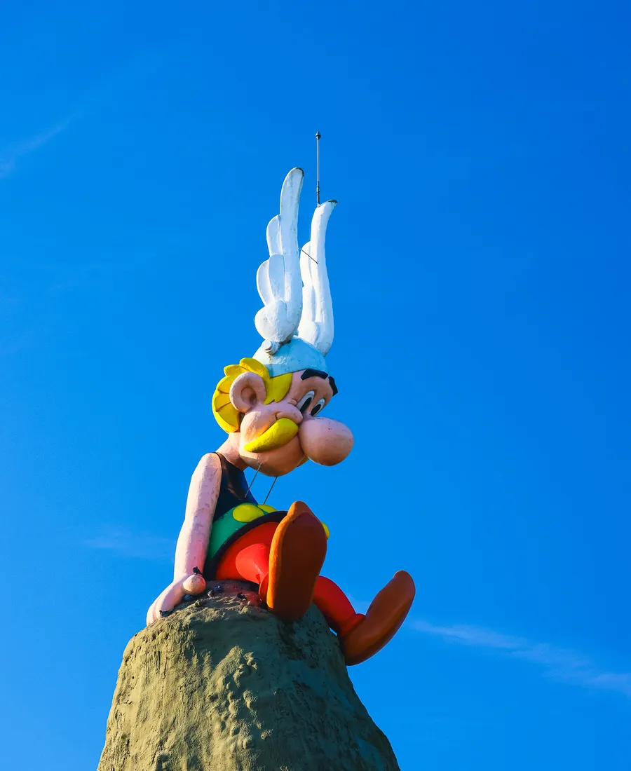 Statue d’Astérix assis au sommet d’un rocher, casque ailé et jambes pendantes, sur fond de ciel bleu