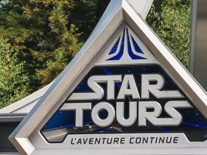 Enseigne triangulaire argentée et bleue de Star Tours L’Aventure Continue devant la végétation, Discoveryland.