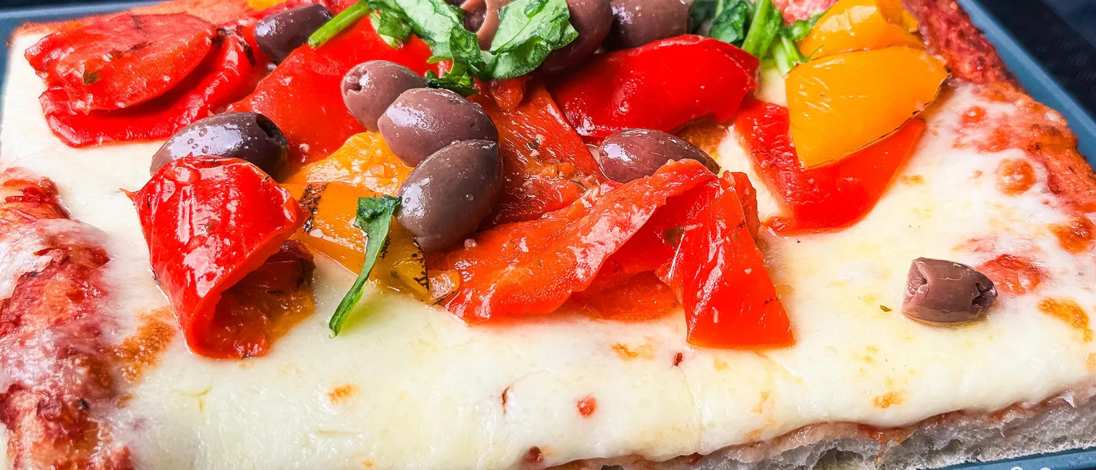 Part de pizza rectangulaire à la mozzarella, poivrons rouges grillés, olives noires et basilic frais.