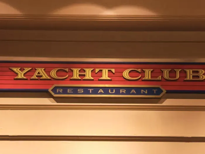 Enseigne en bois rouge et or Yacht Club Restaurant fixée au mur beige, décor maritime du Disney Newport Bay Club.