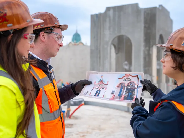 Sur le chantier de la future attraction Les Chaises Volantes des Explorateurs, trois membres du projet en tenue de sécurité examinent un visuel de la façade d’entrée, devant la structure en construction.