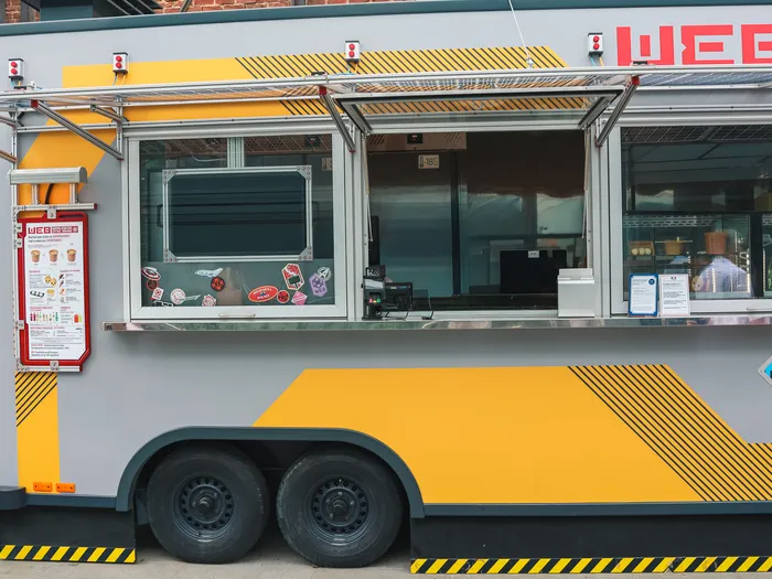 Camion-restaurant WEB Food Truck jaune et gris, comptoir ouvert et menu affiché, dans le parc