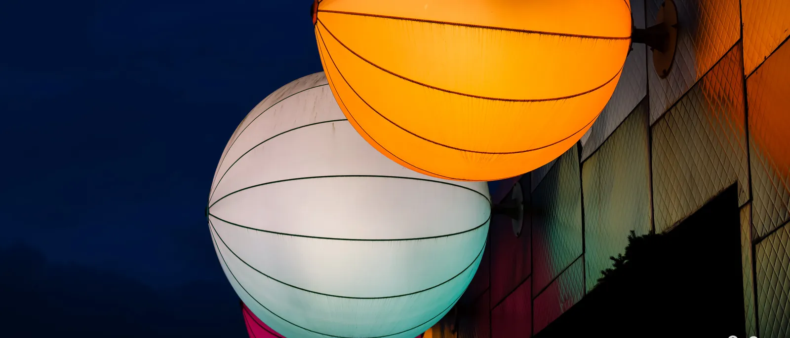 Grosses lanternes sphériques orange, blanche et rose illuminées au crépuscule sur une façade du Disney Village