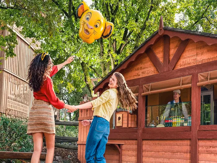 Deux visiteuses souriantes tenant un ballon Simba devant le stand de rafraîchissements en bois Critter Corral, à Frontierland.
