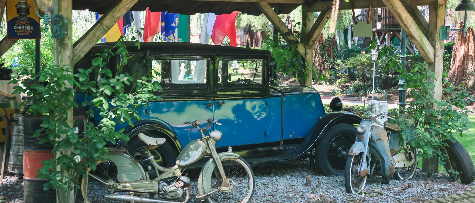 Ancienne voiture bleue et vieille mobylette sous un auvent en bois entourées de plantes à Europa-Park