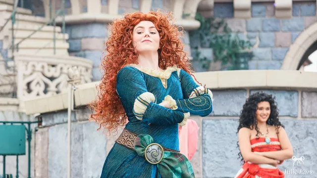 La princesse Mérida, chevelure rousse bouclée et robe bleu-vert, les bras croisés devant le château à Disneyland Paris.