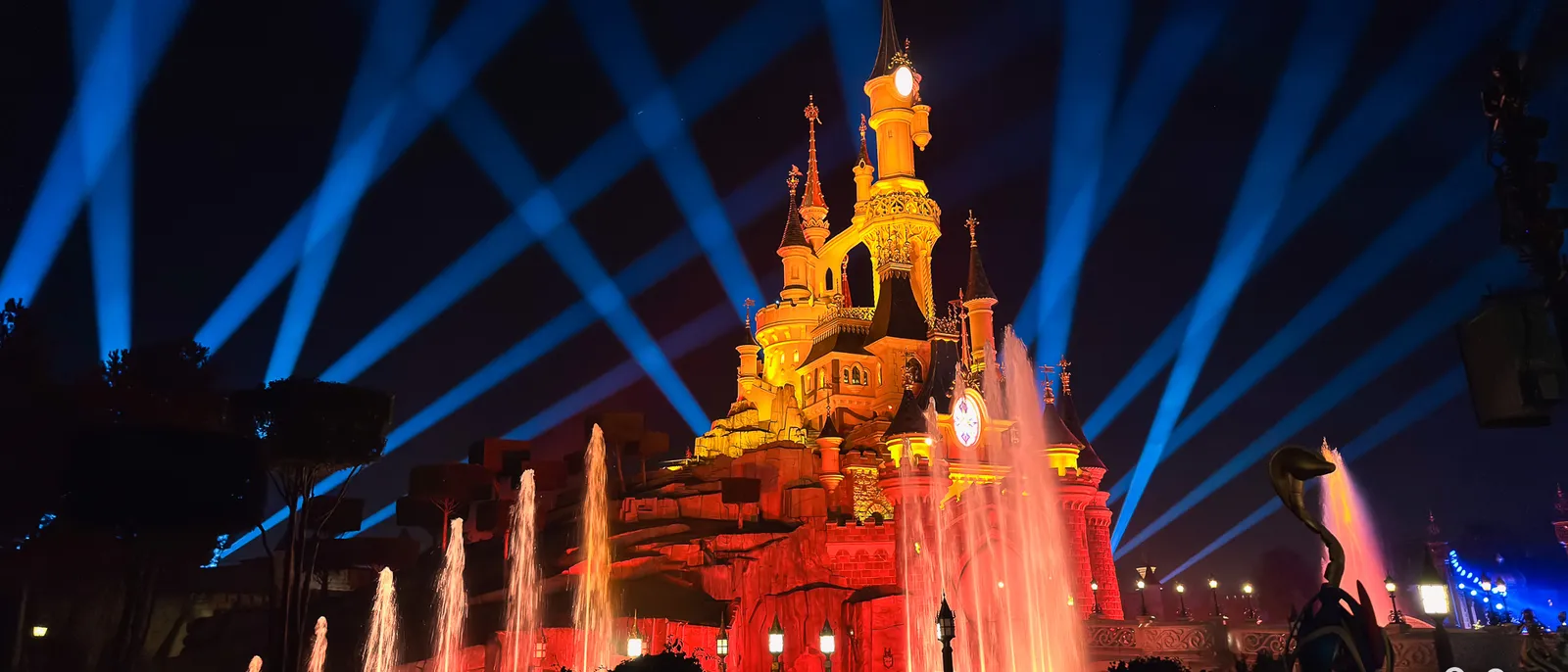 Château illuminé d’orange entouré de jets d’eau et de faisceaux lumineux bleus lors d’un spectacle nocturne.