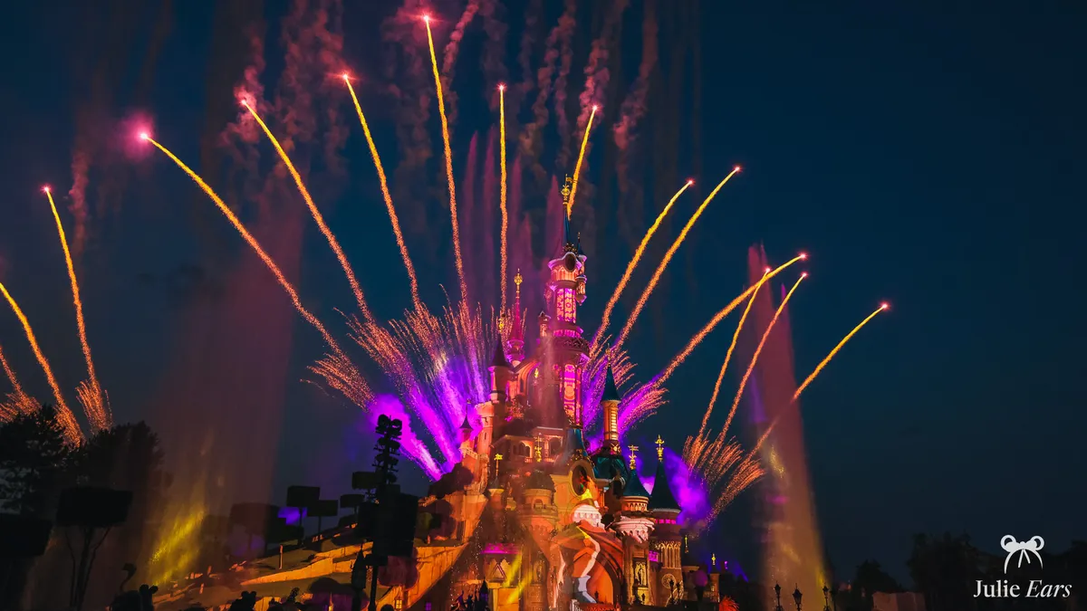 Gold and purple fireworks over Sleeping Beauty Castle during Disney Tales of Magic at Disneyland Paris, with fountains and the audience in silhouette
