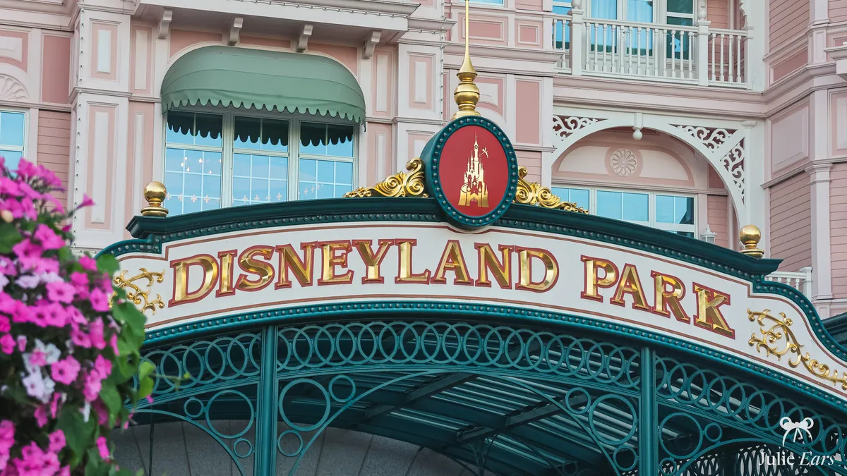 Gold "Disneyland Park" sign on the green wrought-iron entrance arch, in front of the pink Disneyland Hotel, with pink flowers in the foreground