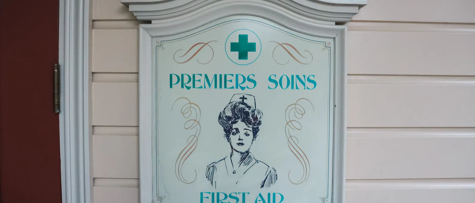 Panneau vintage "Premiers Soins / First Aid" avec croix verte et infirmière dessinée, sur un mur crème à Disneyland Paris