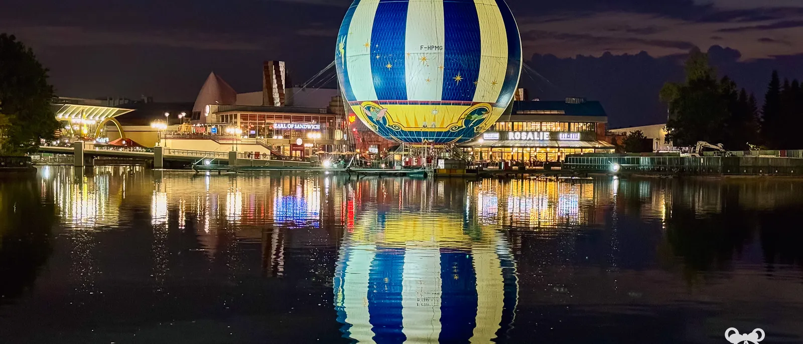 Le ballon PanoraMagique illuminé la nuit au-dessus du lac Disney, reflété dans l’eau, au Disney Village.