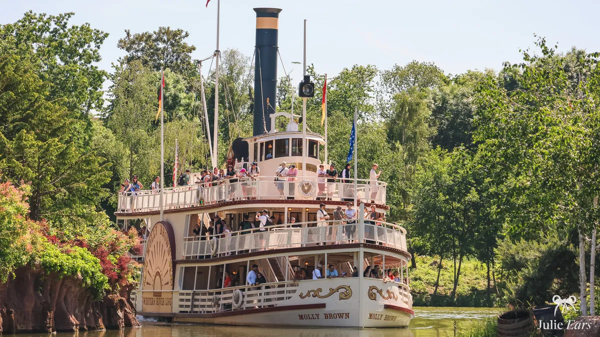 The Molly Brown paddle steamer, its decks full of visitors, sails on the Rivers of the Far West between the trees of Frontierland, at Disneyland Paris